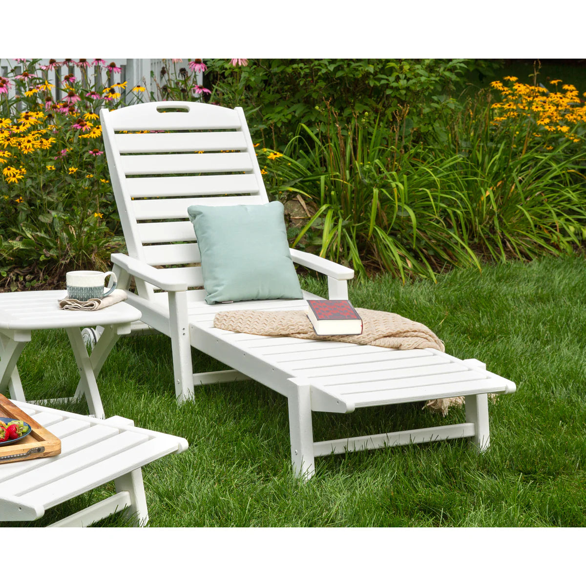 White lounge chair with a pillow and blanket on a grassy area with flowers in the background