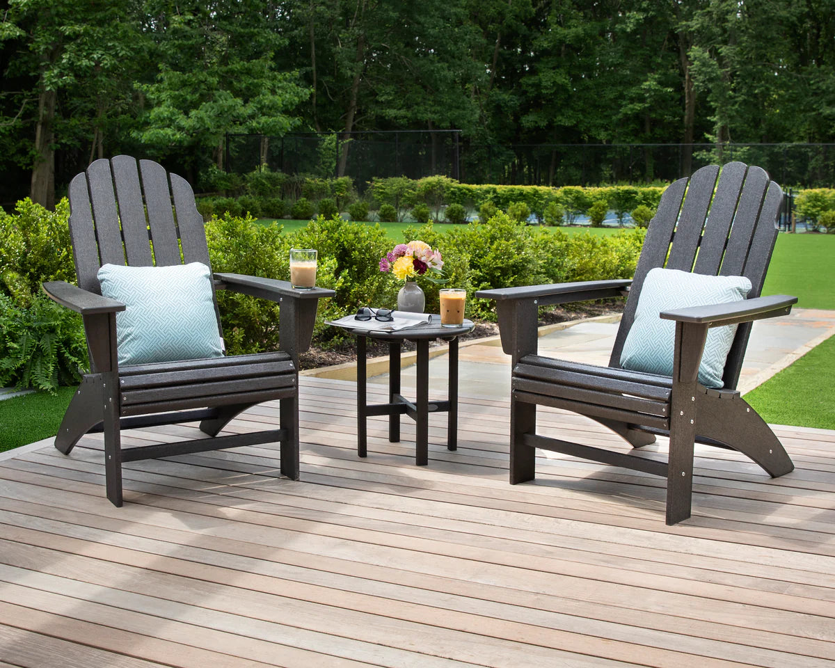 Two Adirondack chairs with cushions on a wooden deck with a small table.