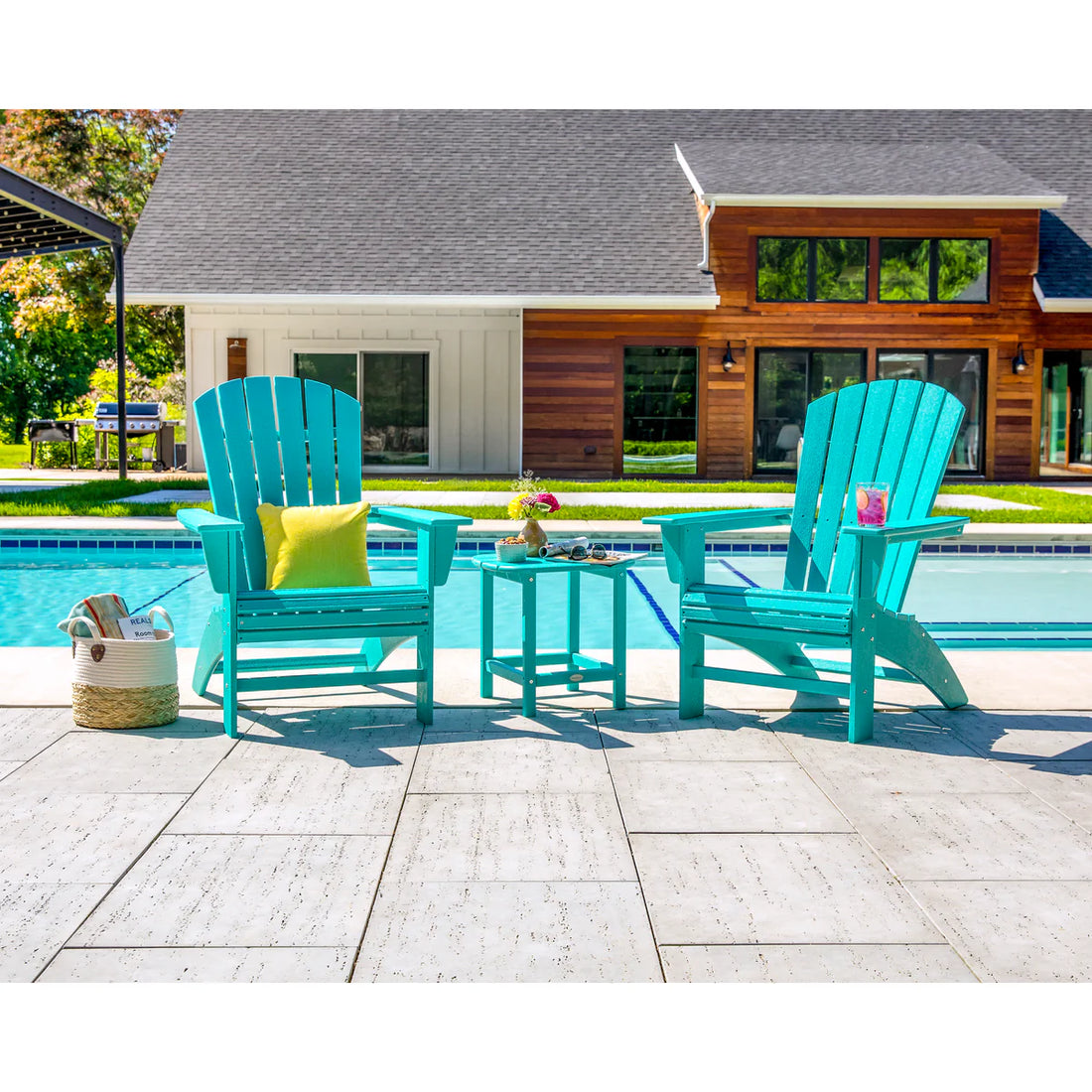 Two turquoise Adirondack chairs with a small table by a poolside, with a house in the background.