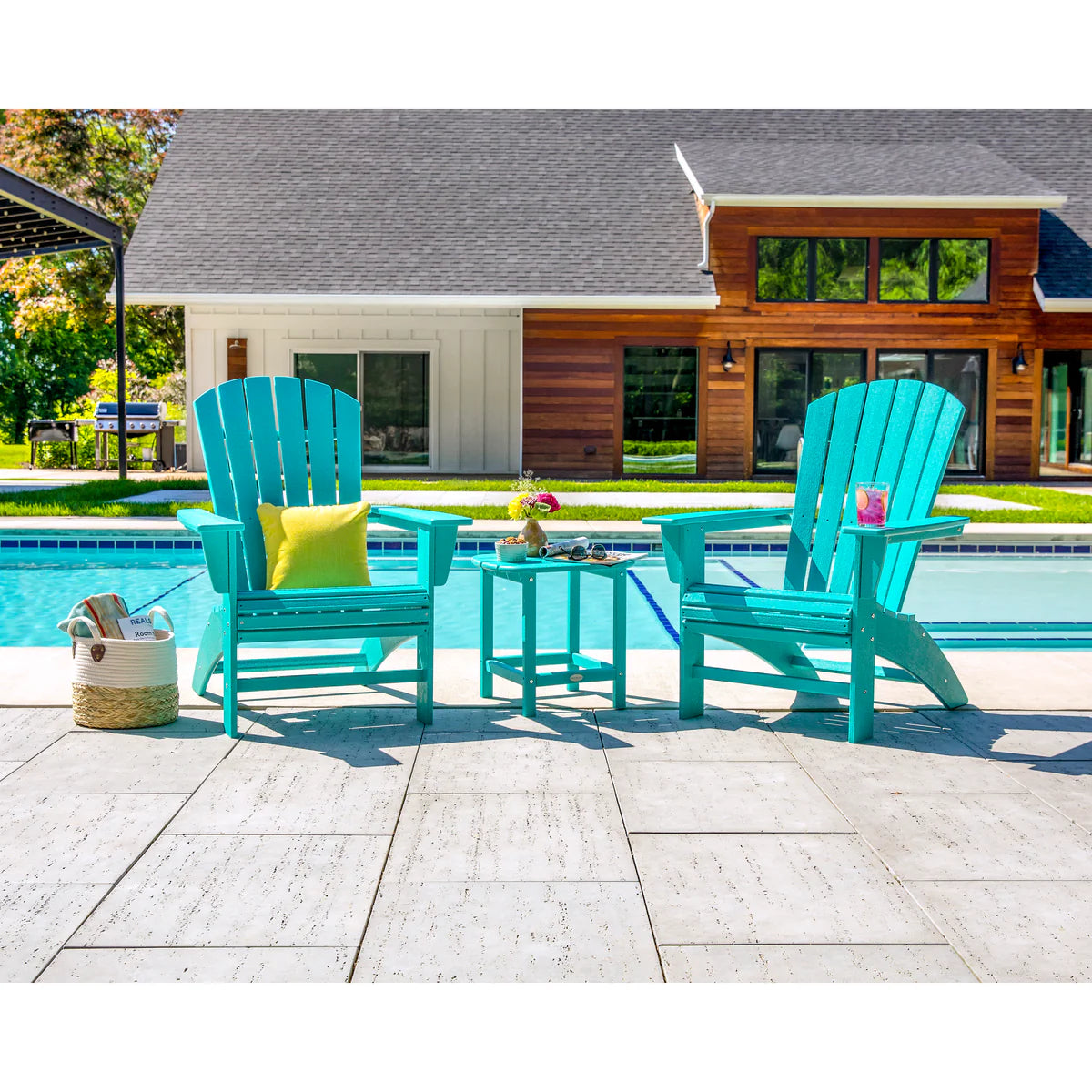 Two turquoise Adirondack chairs with a small table by a poolside, with a house in the background.