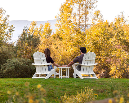 Two people sitting in Adirondack chairs on a grassy area with trees in the background.